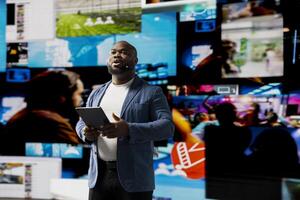 Black presenter hosting a conference on stage surrounded by media visuals, symbolizing a global convention with live audience to discuss about online trends and mobile apps. Public event. photo