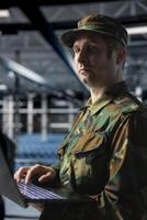 Military officer managing logistics operations using laptop in server room command center. Soldier wearing uniform running diagnostics on infrastructure in army data center photo