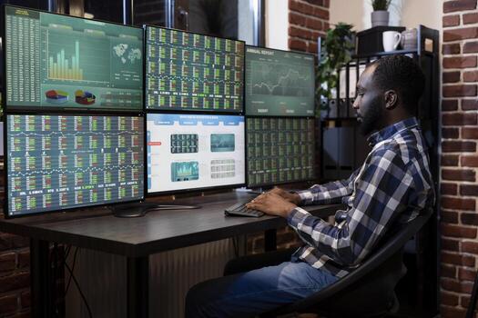 African american trader monitoring forex charts and market trends on multiple screens in brick wall office. Black man types on computer keyboard, analyzing financial data and stock movements. photo