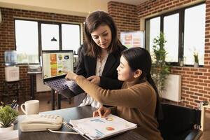 Caucasian employee stands holding laptop, receiving guidance from her asian manager in brick wall office. Young businesswomen using digital device and documents, discussing strategy ideas. photo