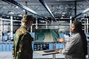 Military server hub personnel discussing target coordinates displayed on digital map interface. Army information expert talking with commander in data center, reviewing live tactical feed on screen photo