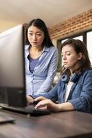 Closeup of caucasian employee typing on keyboard as asian manager stands and reviews the work on desktop monitor. Two professional women using computer, having a creative discussion in modern office. photo