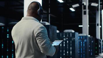 Admin walking in data center, listening music while doing inspection on infrastructure tech. IT specialist in server hub wearing headphones analyzing traffic patterns of equipment mainframes, camera B photo