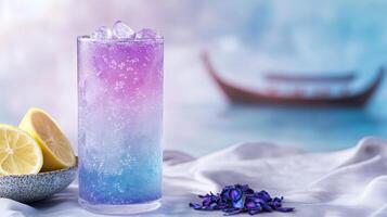 A colorful cocktail with purple and blue hues is elegantly presented beside fresh lemon slices and dried flowers near a calm body of water photo
