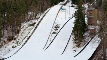 An aerial view shows a ski jumping ramp covered in fresh snow, surrounded by trees in a serene winter landscape photo