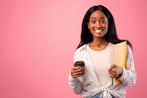 Smiling African American woman with notebooks and takeaway coffee on pink studio background. Portrait of black female student looking at camera, ready to start school. Education concept photo