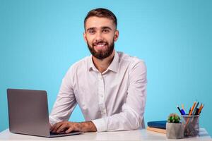 Millennial businessman using laptop at his desk, working online in office, blue studio background. Smiling young entrepreneur working with computer, browsing web, participating in online conference photo