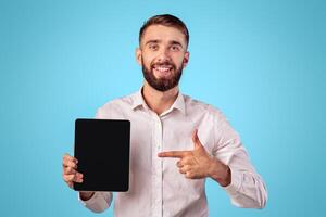 Young businessman pointing at tablet with empty screen on blue studio background, mockup for application or website design. Cheerful millennial guy showing touch pad with space for your ad photo