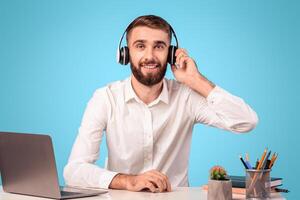Bearded male helpline operator with headphones and laptop answering customer's call at desk over blue studio background. Happy tech support manager communicating with client photo