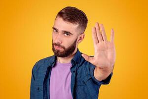 Serious hipster guy gesturing STOP, showing palm to camera over grey studio background. Young man expressing his negative attitude, rejecting or prohibiting something photo