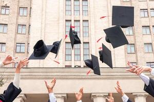Multiracial group of students standing in a row and throwing graduation caps up in the air, posing next to university building after graduation ceremony, celebrating freedom, panorama with copy space photo