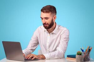 Online job concept. Attractive young businessman using laptop computer at his desk on blue studio background. Focused millennial office employee working on pc, typing, browsing internet photo