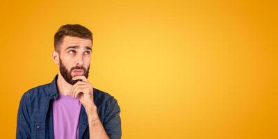 Portrait of pensive young bearded man touching his chin, deep in thought over orange studio background. Doubtful millennial guy thinking and making decision, dreaming about something photo