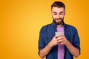 Handsome bearded guy with smartphone browsing internet, communicating online or shopping over orange background. Young man working or studying remotely using mobile device photo