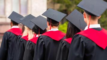 Back view of international group of students in graduation robes and caps walking by university campus, closeup. Unrecognizable multiracial students attending graduation ceremony photo