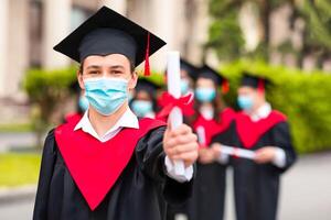 Caucasian guy student in graduation costume and protective face mask showing his diploma at camera, posing over international group of students at university campus, selective focus, copy space photo