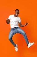 Cute black guy winner jumping up in the air and showing funny figures, having good day, showing positive emotions over orange background. African american man posing at studio, having fun photo