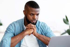 Portrait Of Thoughtful Black Freelancer Guy Sitting At Desk And Looking At Laptop Screen, Pensive African Man Thinking About Business Ideas While Working On Computer At Home Office, Free Space photo