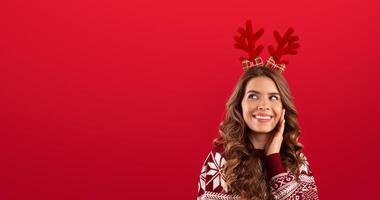 It's Christmas time. Beautiful young lady in colorful warm sweater wearing deer horns on red background. Panorama photo