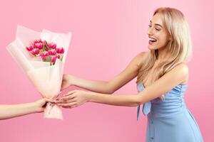 Joyful young lady receiving bouquet of beautiful spring flowers for Woman's Day on pink studio background. Millennial blonde feeling happy to get lovely tulips as holiday gift photo
