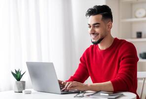 Handsome Arab Male Freelancer Working On Laptop At Home Office, Smiling Young Middle Eastern Man Sitting At Desk With Computer In Light Room And Typing On Keyboard, Enjoying Remote Work, Copy Space photo