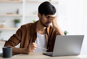 Tired indian man sitting in front of laptop at office, touching his head, suffering from burnout or financial crisis, young guy freelancer having difficulties with job, lack of clients, copy space photo