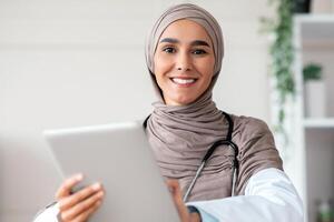 Smiling woman in hijab with stethoscope doctor using digital tablet, sitting at workdesk in clinic, copy space. Female doctor using modern technologies for communication with patients, telemedicine photo