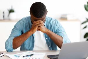 Depressed African American Entrepreneur Having Problems At Work. Stressed Black Businessman Sitting At Desk With Laptop Computer And Touching Head In Despair, Suffering Bankruptcy, Free Space photo