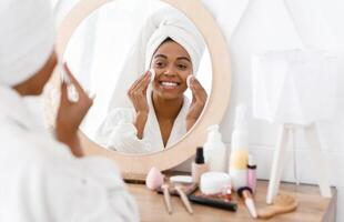 Pretty african american woman removing makeup, cleaning face with cotton pads, looking in mirror while sitting at dressing table photo