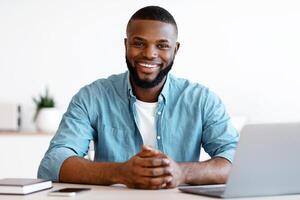 Portrait Of Positive African American Freelancer Man Sitting At Desk In Home Office. Handsome Black Guy Self-Entrepreneur Working At Table With Laptop Computer, Looking At Camera And Smiling photo