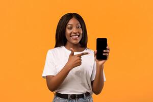 Smiling young black lady showing modern mobile phone, pointing at empty screen, sharing her positive emotions after using nice mobile application, orange studio background, copy space photo