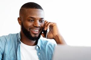 Closeup Shot Of Cheerful African American Guy Talking On Cellphone And Working On Laptop At Home Office, Smiling Black Freelancer Man Looking At Computer Screen, Enjoying Remote Work, Copy Space photo