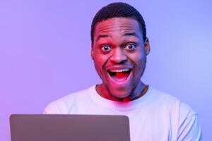 Online Offer. Closeup Portrait Of Of Joyful Young African Man Using Laptop Computer And Looking At Camera With Excitement, Happy Black Guy Standing Under Neon Light Over Purple Background, Free Space photo