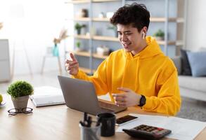Online Education, Remote Work. Smiling asian man having videochat with colleagues or class group of students, using laptop. Guy wearing wireless earbuds, talking to webcam, explaining and gesturing photo