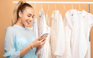 Fashion And Style Concept. Portrait of smiling female customer holding and using mobile phone, texting sms or making purchases in online store standing near rack with clothes at pink studio photo