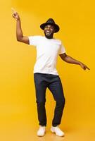 Cheerful black man in hat dancing and pointing index fingers aside on yellow studio background photo