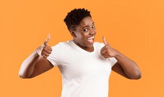 Funny Plus-Size Black Lady Gesturing Thumbs Up With Both Hands Smiling To Camera And Approving Something Standing Over Yellow Studio Background. Thumbs-Up, I Like It Gesture Concept photo