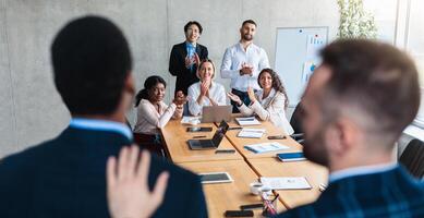 Manager Introducing New Male Worker To Business Team During Corporate Meeting Standing In Modern Office. Rear View, Selective Focus On Diverse Colleagues Sitting At Desk. Employment And Recruitment photo