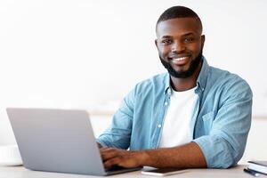 Portrait Of Happy Successful Black Freelancer Guy Posing At Desk With Laptop, Positive Millenial African American Man Smiling At Camera While Working Online On Computer In Home Office, Free Space photo