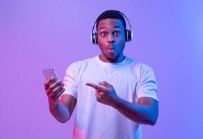 Cool App. Surprised Black Guy In Wireless Headphones Pointing At Smartphone In Hand, Emotionally Reacting To New Mobile Application Or Music Streaming Service, Standing Under Neon Light, Copy Space photo