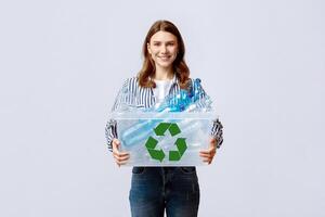 Waste Reuse. Cheerful Young Woman Carrying Container With Plastic Bottles For Recycling And Smiling At Camera, Happy Eco-Activist Sorting Garbage For Recycle, Standing On Grey Background photo