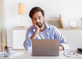 Overworked arab male entrepreneur sitting at desk in office and sleeping, eastern man napping at workplace, placed head on hand, feeling tired after working on laptop computer, copy space photo