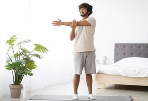 Cheerful millennial indian man in homewear practicing yoga at home, standing on fitness mat and stretching arms, looking at copy space and smiling, enjoying morning workout, panorama photo