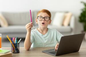 Inspired school kis redhead boy with glasses using laptop at home, raising hand with pencil up, smart preteen child showing eureka gesture, looking for creative solutions while studying, copy space photo