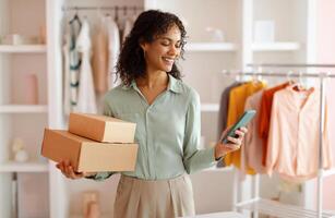 Smiling entrepreneur carrying cardboard boxes while checking her smartphone in a modern fashion studio with a rack of clothes in the background photo