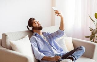 Domestic Relax. Young Arab Man In Wireless Headphones Listening Music And Browsing Internet On Smartphone While Resting On Comfortable Sofa At Home, Relaxing In Cozy Living Room, Free Space photo