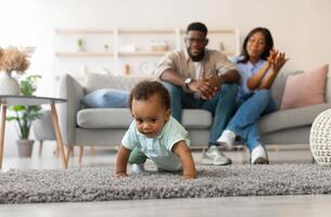 Portrait of adorable cute baby boy learning how to walk at home, crawling on rug floor carpet, happy mom and daddy proud of their little child sitting on sofa in blurred background, mum clapping hands photo