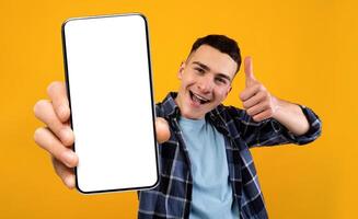 Mobile Application Ad. Excited Young Guy Showing Smartphone With Blank White Screen And Gesturing Thumb Up, Cheerful Man Recommending New App While Standing Over Yellow Studio Background, Mockup photo