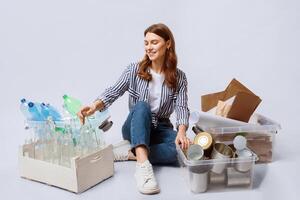 Portrait Of Young Woman Sitting Among Containers With Sorted Garbage, Millennial Smiling Lady Enjoying Zero Waste Living And Garbage Recycle, Posing Over Light Background In Studio, Free Space photo
