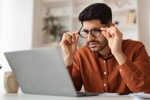 Error. Portrait of confused Arab guy sitting at desk using laptop, taking off glasses looking at pc screen and squinting. Worried man reading bad negative news, having poor eyesight problems photo